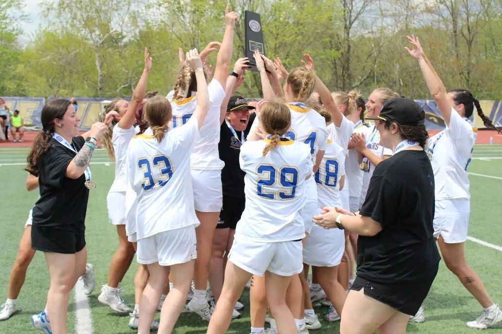 Head coach Kelly Downs holds up the MASCAC trophy as the team gathers around her in celebration after the game (photo credit: Krystyanna Ramsdell)